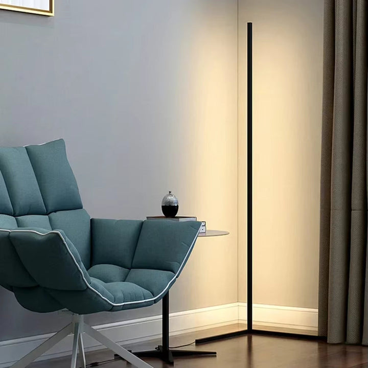 Black corner floor lamp glowing warm white beside a teal padded armchair and a small side table with a decorative vase. 1