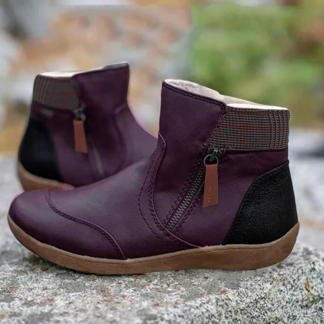 Pair of dark purple ankle boots with side zipper, brown pull tab, plaid collar and black heel panel, set on a stone.