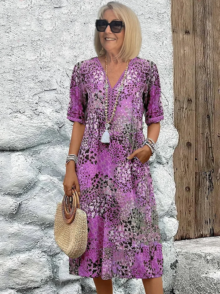 Model posing in the V-neck midi dress in a purple speckled print, with tasseled beaded necklace and round straw bag. 4