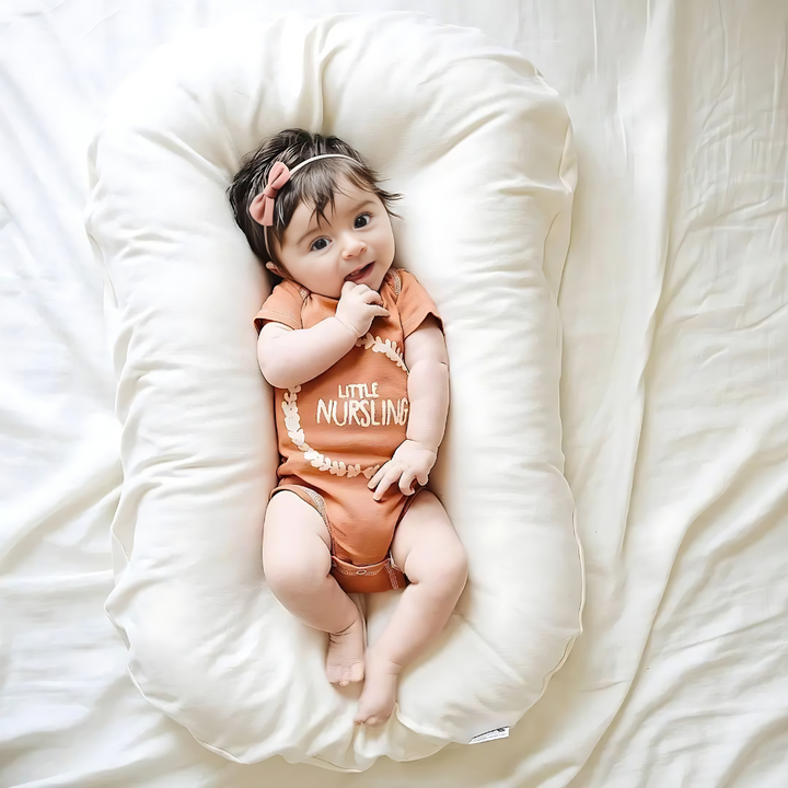 Overhead view of a baby with a pink bow resting in a cream cushion on white bedding 5