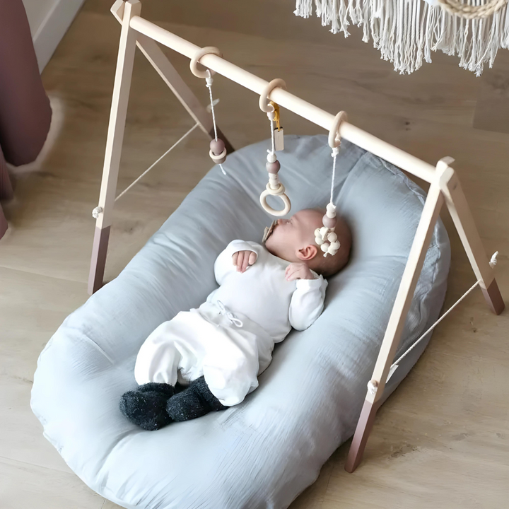 Baby lying on a light blue-gray pillow beneath a wooden play gym with hanging toys 6