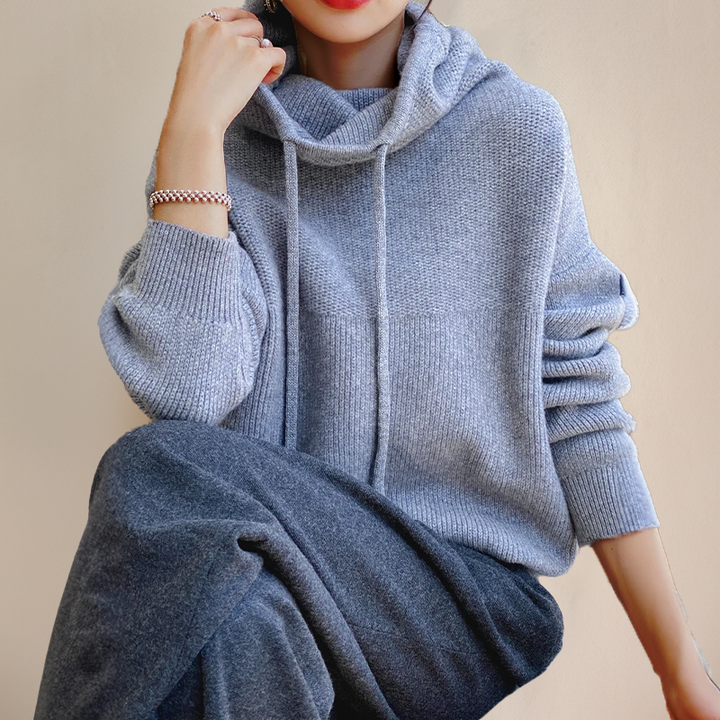 Seated model in a light gray drawstring cowl-neck sweater with ribbed cuffs, dark gray trousers and a beaded bracelet 5