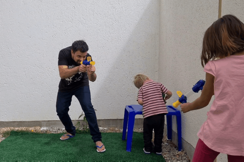 Man and a girl aim blasters in a small yard while a toddler stands at a blue stool 10