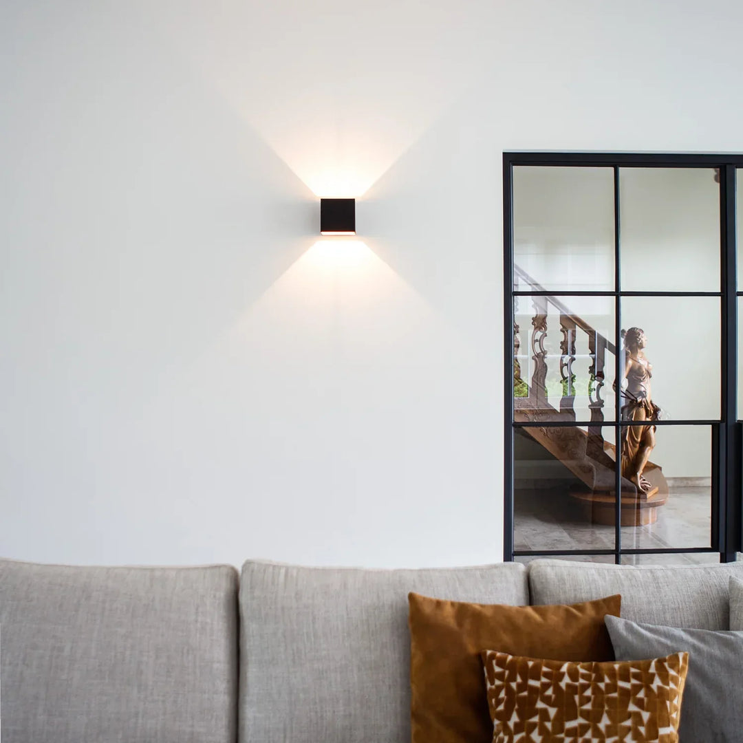 Black cube wall lamp on a white wall above a gray sofa with orange cushions, next to a black-framed glass door. 10