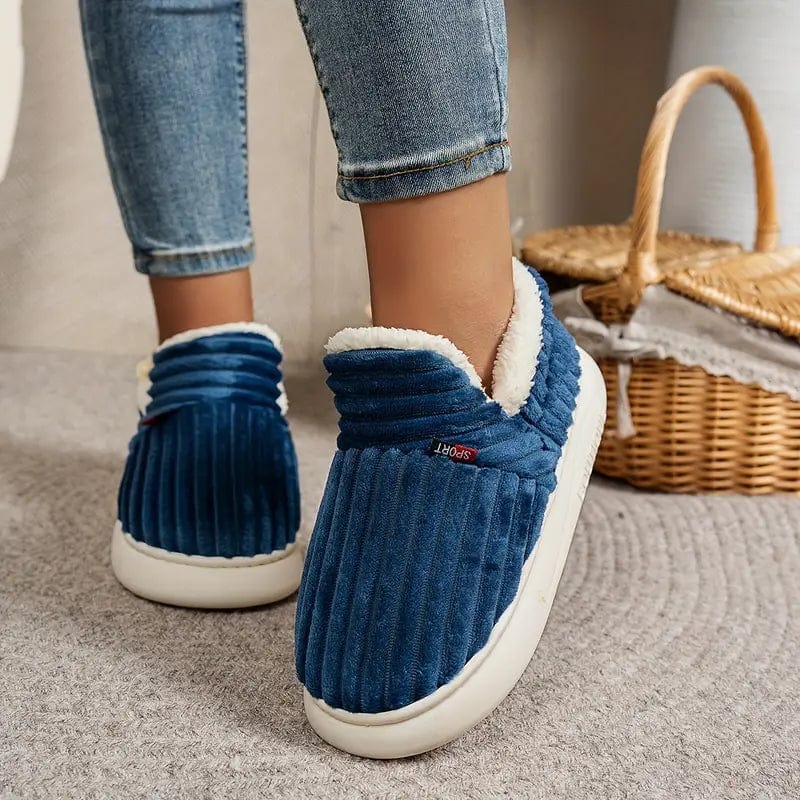 Person wearing blue ribbed fleece-lined slippers with jeans, standing on a gray rug beside a wicker basket.