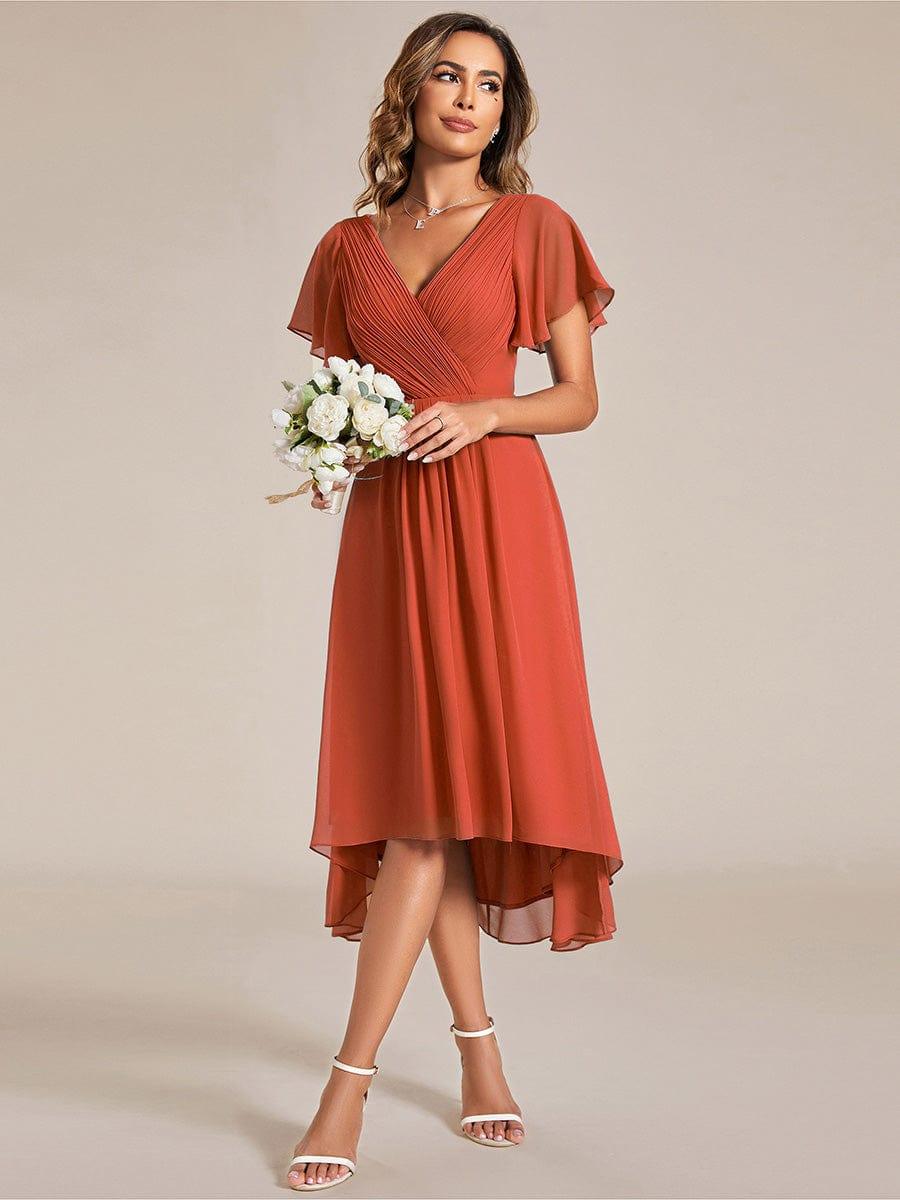 Model in the burnt orange chiffon dress holding a bouquet of white flowers, glancing to the side in white strappy heels. 95
