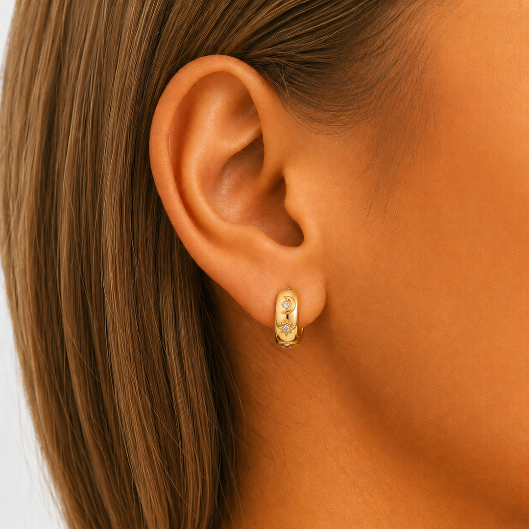 Close-up of a gold huggie hoop worn in an ear, with a small moon and star motif set with tiny clear stones. 3
