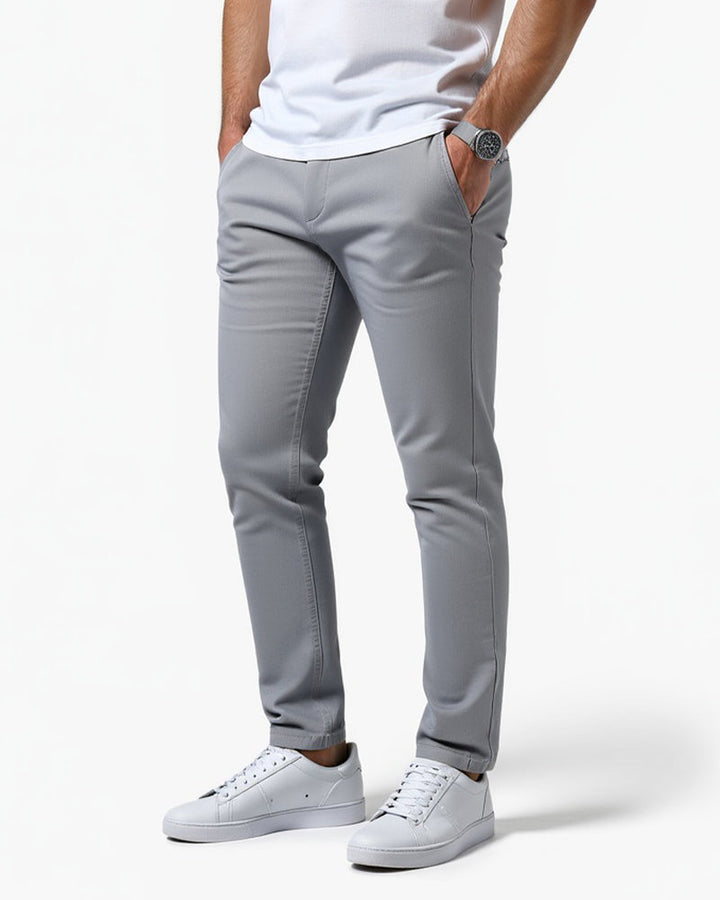 Light gray slim chinos seen from a three-quarter front angle, worn with a white tee, white sneakers and a wristwatch, one hand in pocket. 1