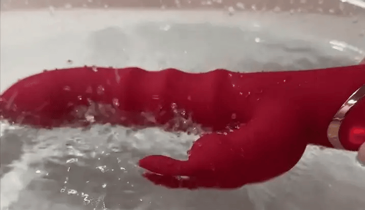 Red silicone massager with a ribbed shaft and curved arm submerged in splashing water, demonstrating its waterproof design. 6