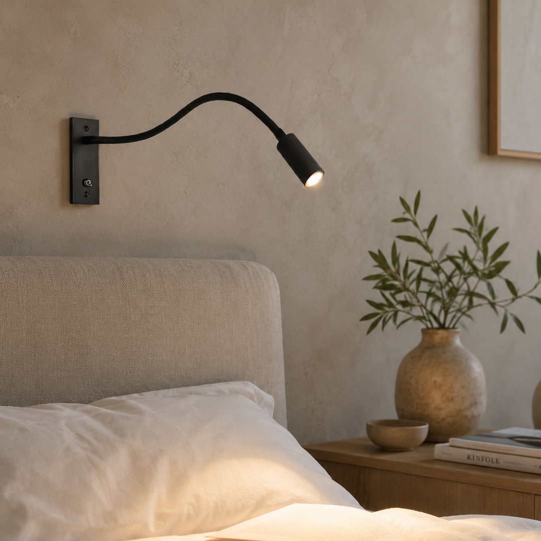 Black gooseneck wall light glowing above a bed with white linen pillows and a nightstand with a vase of olive branches.