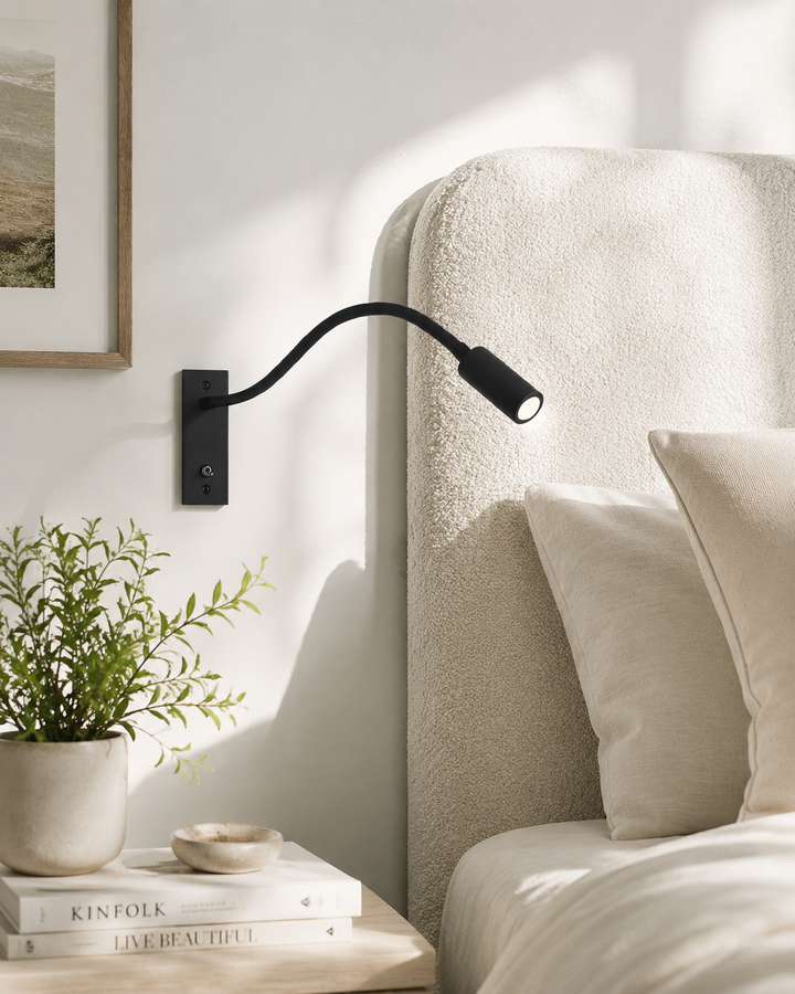 Black gooseneck wall light mounted beside a cream bouclé headboard with a plant and books on the nightstand.