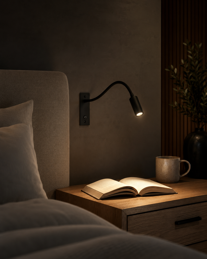 Black gooseneck wall light shining on an open book on a wooden nightstand with a ceramic mug.