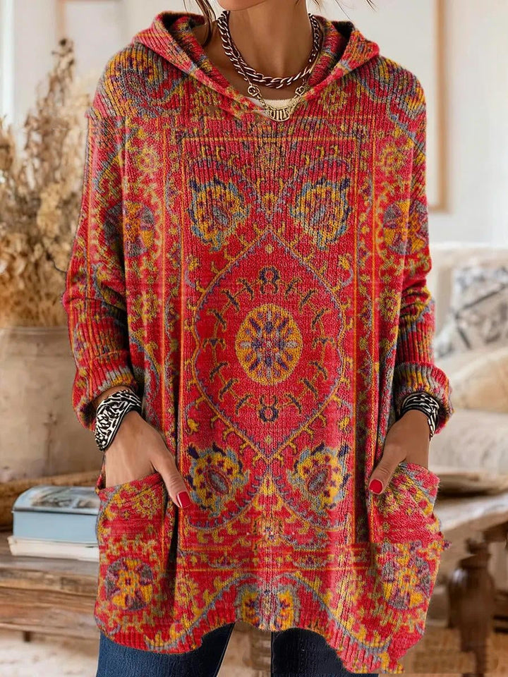 Model wearing the hooded tunic in red with a gold and blue rug-inspired medallion pattern, long sleeves, hands in the pockets. 4