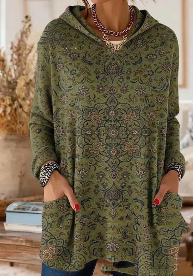 Model wearing the hooded tunic in olive green with a fine navy and tan floral mandala pattern, hands in the front pockets. 7