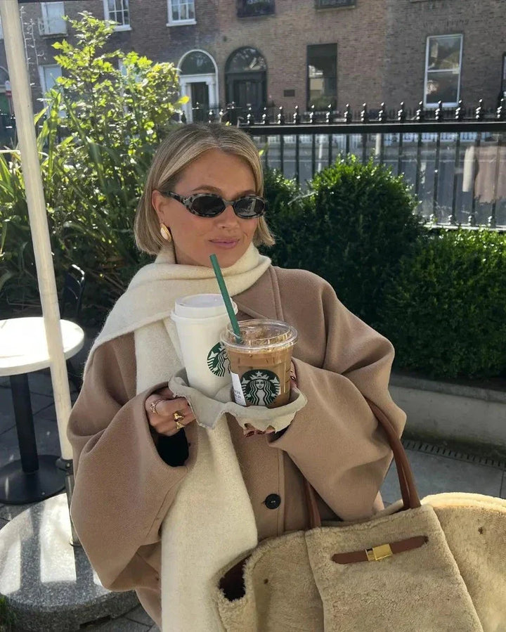 Frau mit Sonnenbrille hält Starbucks-Getränke, trägt beigen Mantel und weiße Schal.