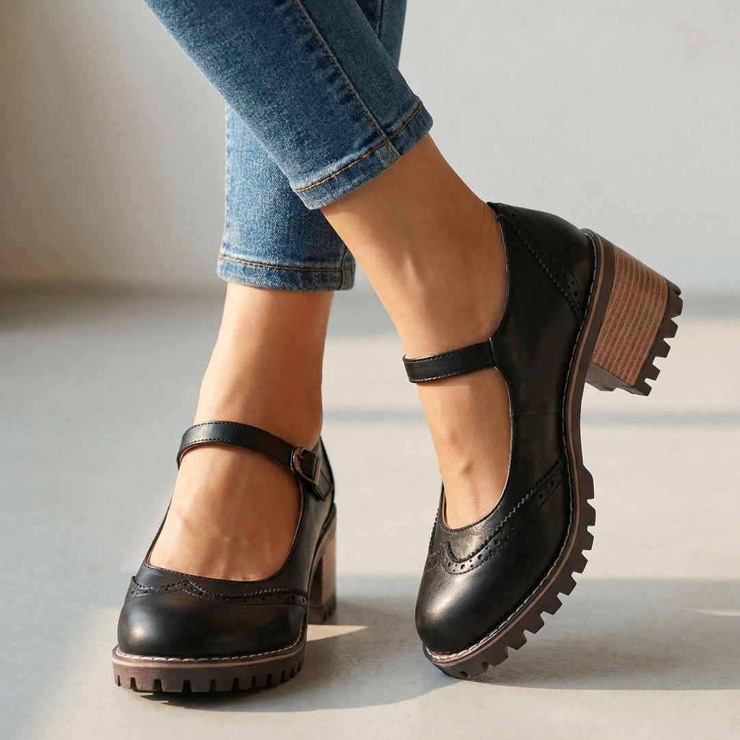 Black Mary Janes with brogue detail, chunky stacked heel, and ankle strap on a plain indoor floor.