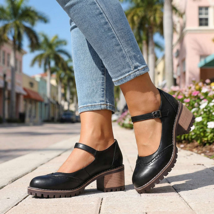 Black pair worn on a sidewalk beside flowers, showing the buckle on the outer side and chunky heel 7