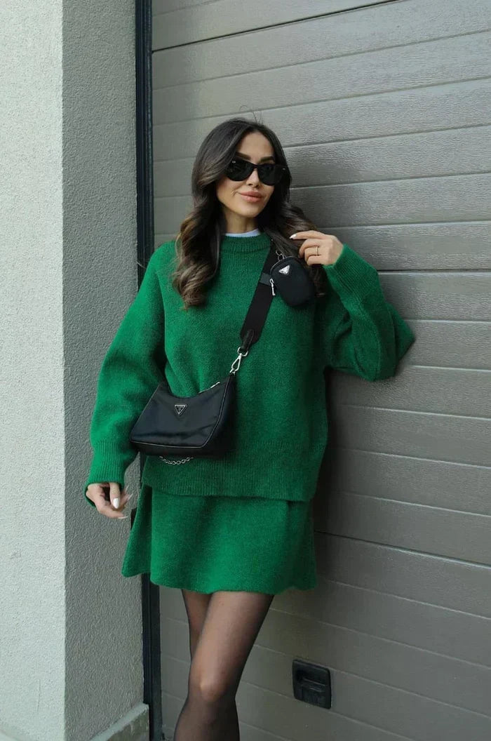 Model leaning on a garage door in the green sweater and skirt set, hand raised to her shoulder 10