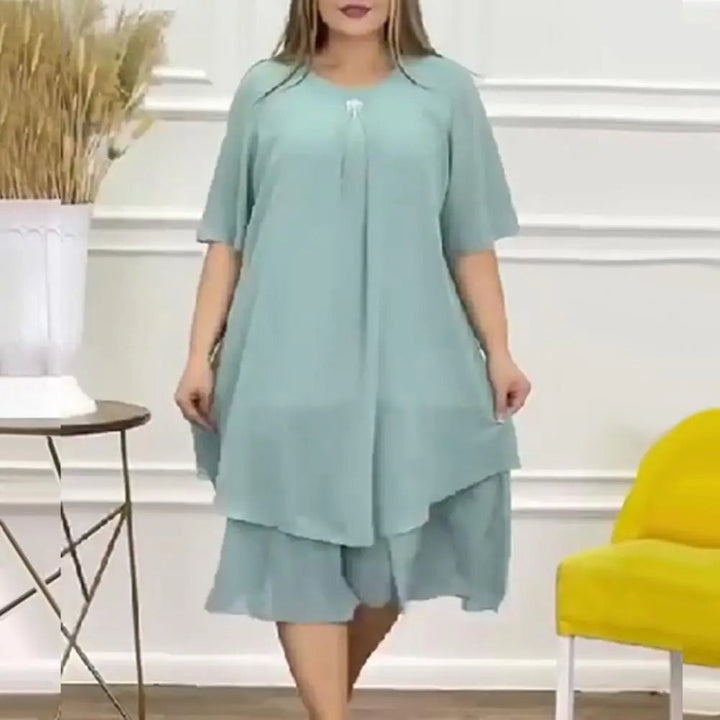 Frau in mintgrünem Sommerkleid aus Chiffon, knielang, mit kurzen Ärmeln, vor weißer Wand.