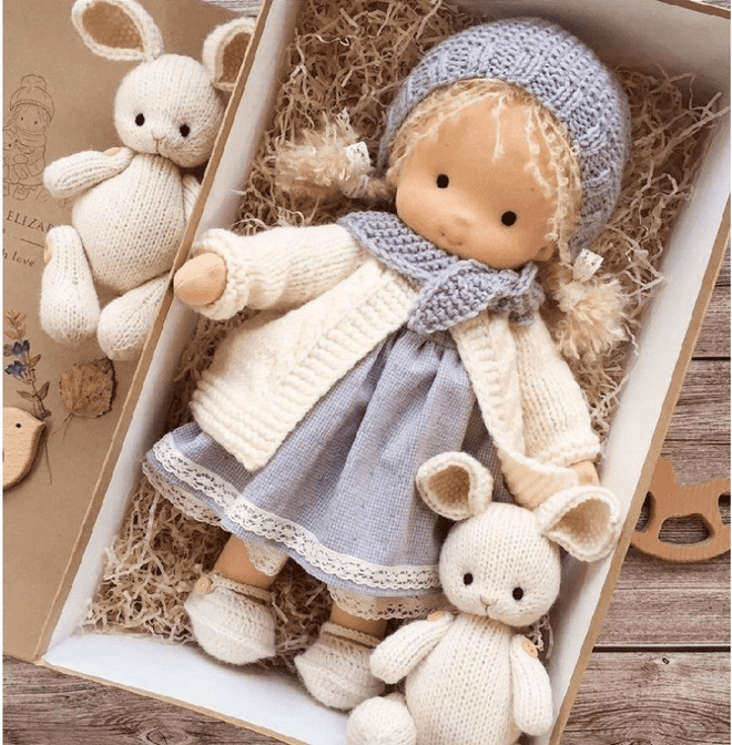 Doll with blue knit bonnet and scarf, cream cardigan and gray dress in a box with two knit bunnies 4