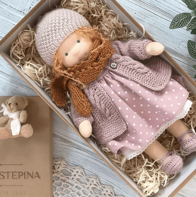 Doll in pink knit hat and cardigan, polka-dot dress and rust scarf lies in a straw-filled box 5