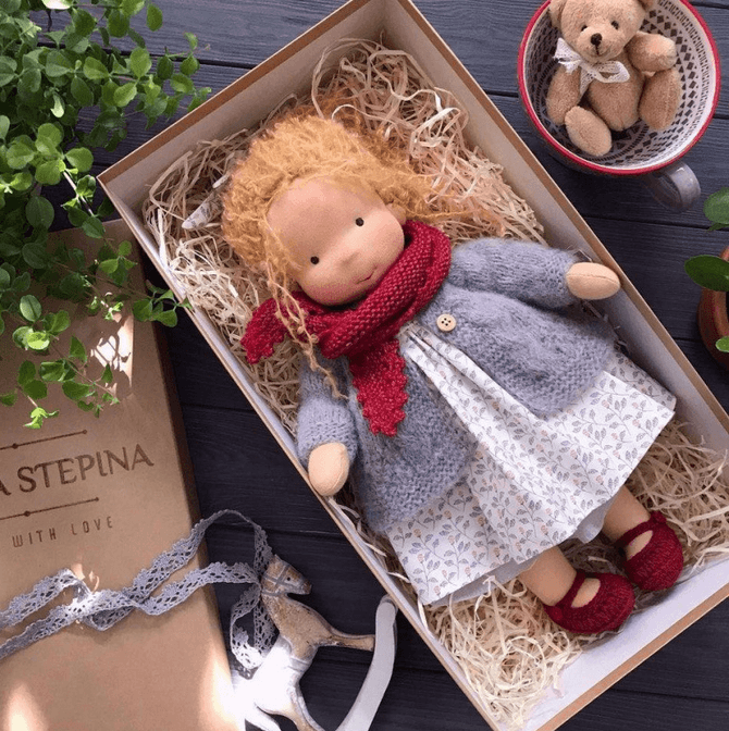 Doll with red-gold curls, red scarf, gray cardigan and floral dress lying in a box with a teddy in a cup 9