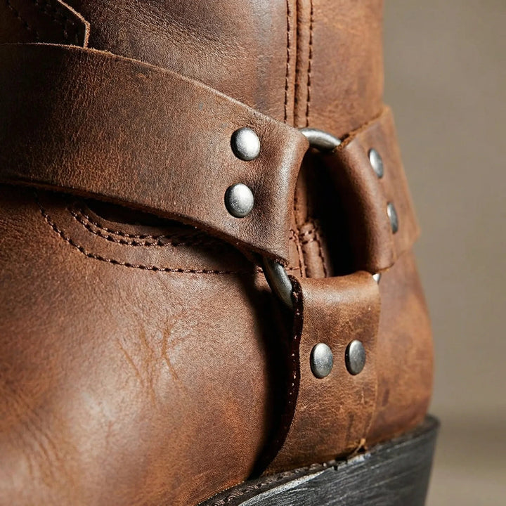 Close-up of the harness strap on a brown leather boot, showing the metal ring, rivets, stitching and grainy leather texture. 3