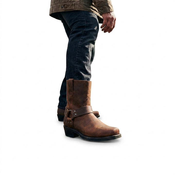 Man in dark jeans and an olive jacket walking in a brown leather harness boot, shown from the knee down. 1