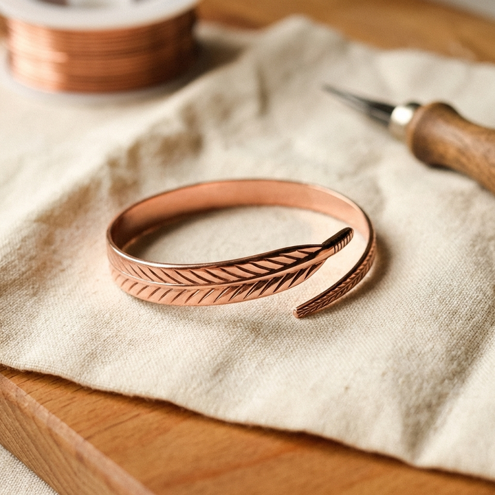 Copper bracelet with a feather engraving wrapping around, resting on linen beside a wire spool and tool 1