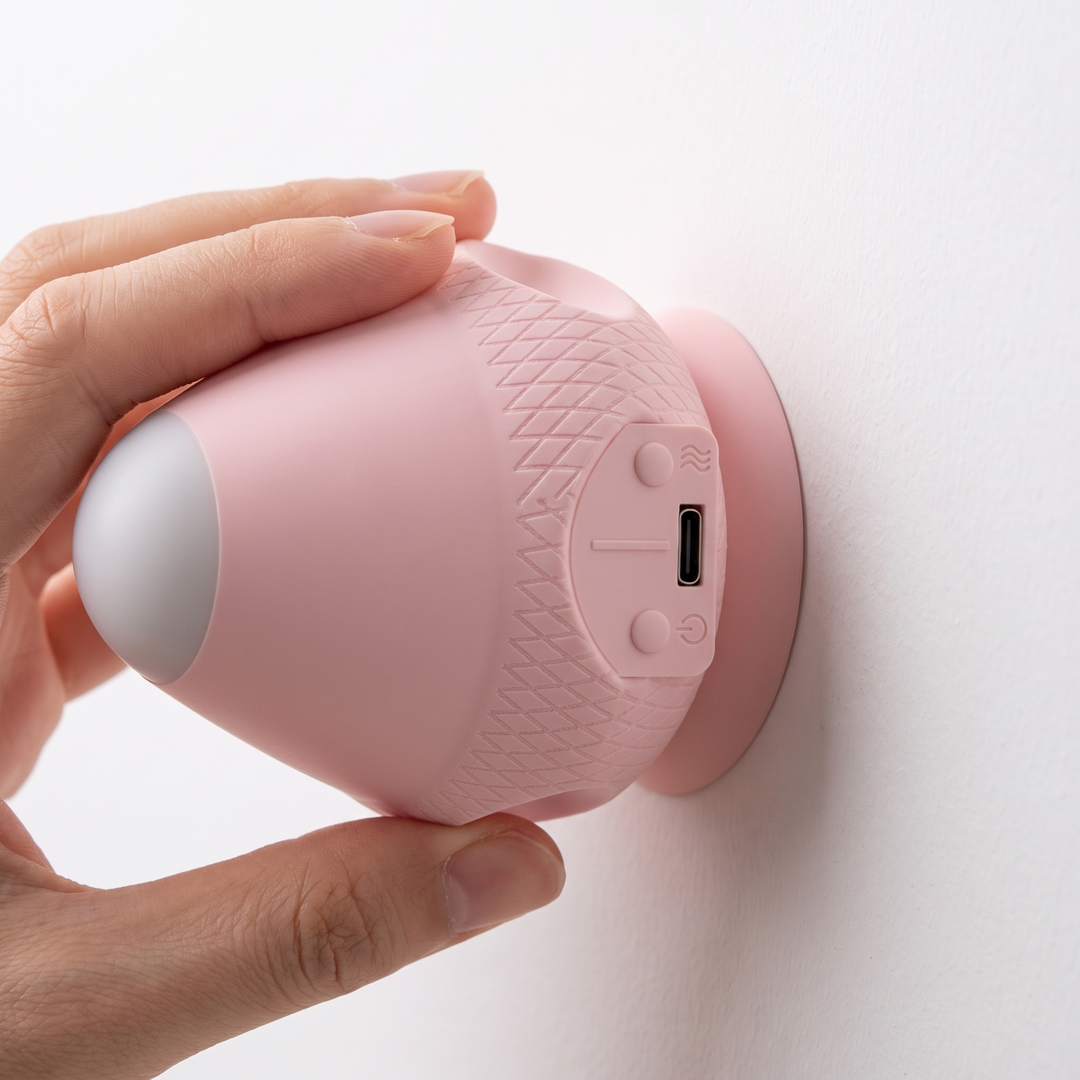 Hand holds a pink massager against a white wall, its suction base attached, with the control buttons and USB-C port visible on the side. 12