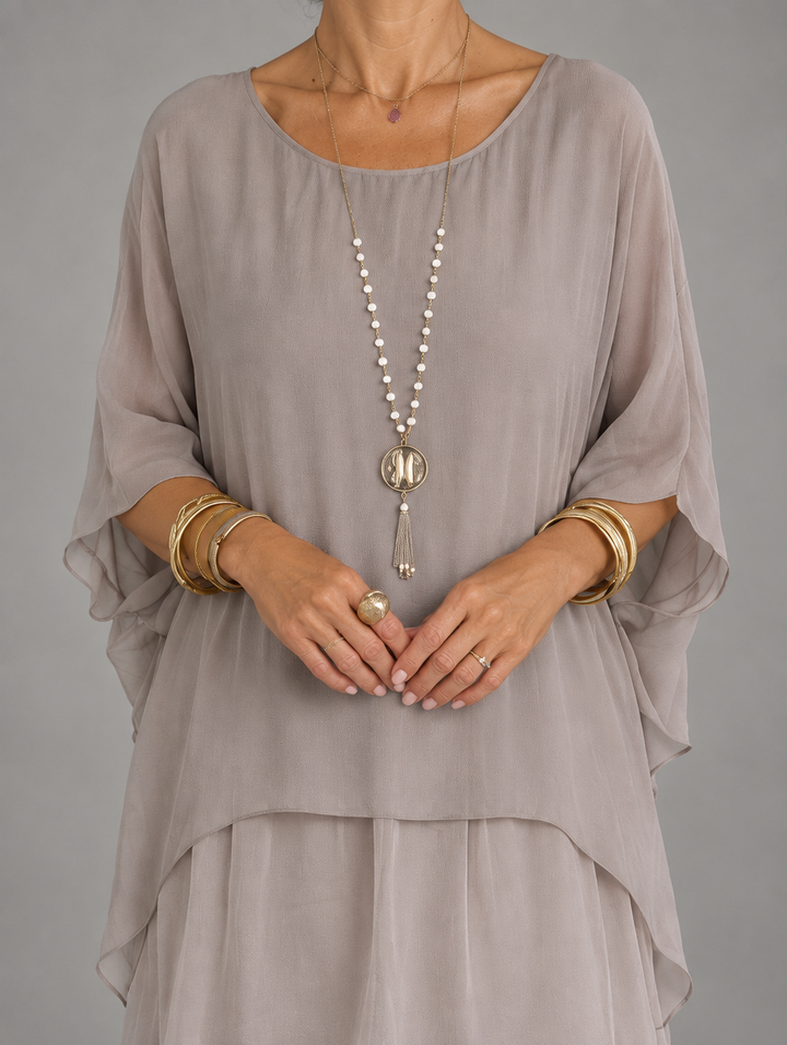 Close-up of a model wearing the taupe-gray version, showing the sheer caped overlay, scoop neckline and layered hem with gold jewelry. 21
