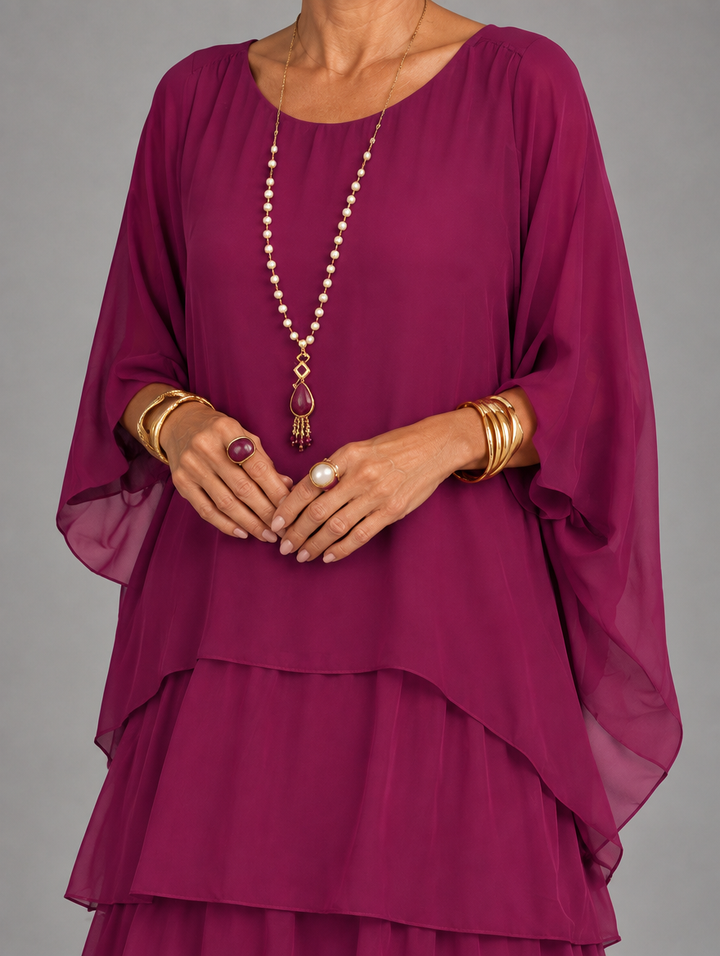 Torso close-up of the berry-colored dress, showing the scoop neckline, flowing cape sleeves and layered overlay with pearl and gold jewelry. 24