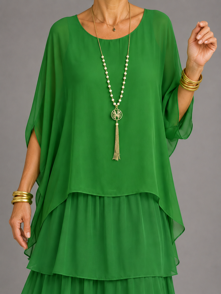 Torso close-up of the emerald green dress, showing the sheer cape sleeves and layered hem with a long beaded tassel necklace. 33