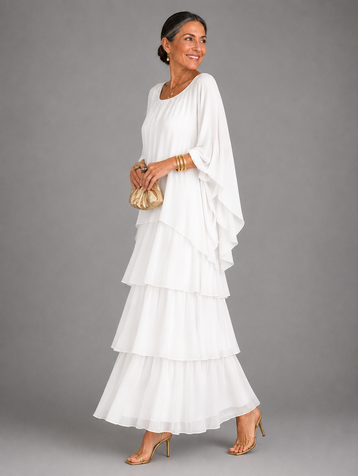 Side view of a model in the white version, with draped cape sleeve and three-tier ruffle skirt, holding a small gold clutch. 28
