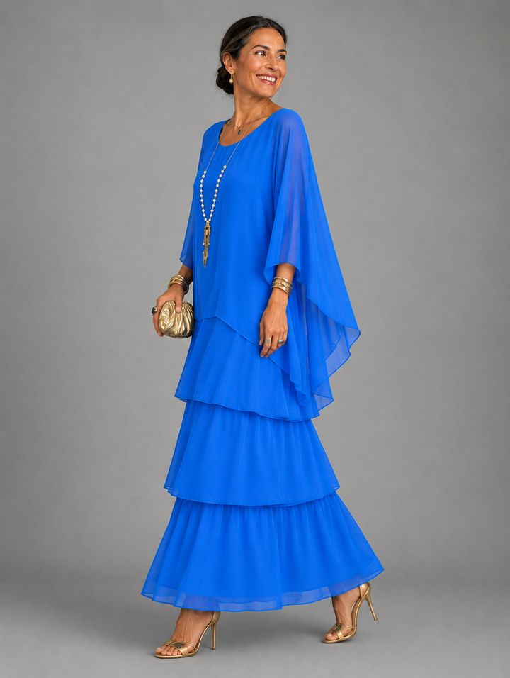 Side view of a model in the bright royal blue dress, with cape overlay and tiered skirt, holding a gold clutch. 25