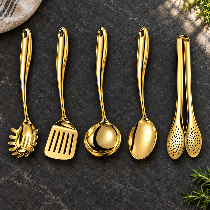 Five gold-colored utensils in a row on dark stone: pasta server, slotted turner, ladle, solid spoon and perforated tongs, with herbs nearby. 24