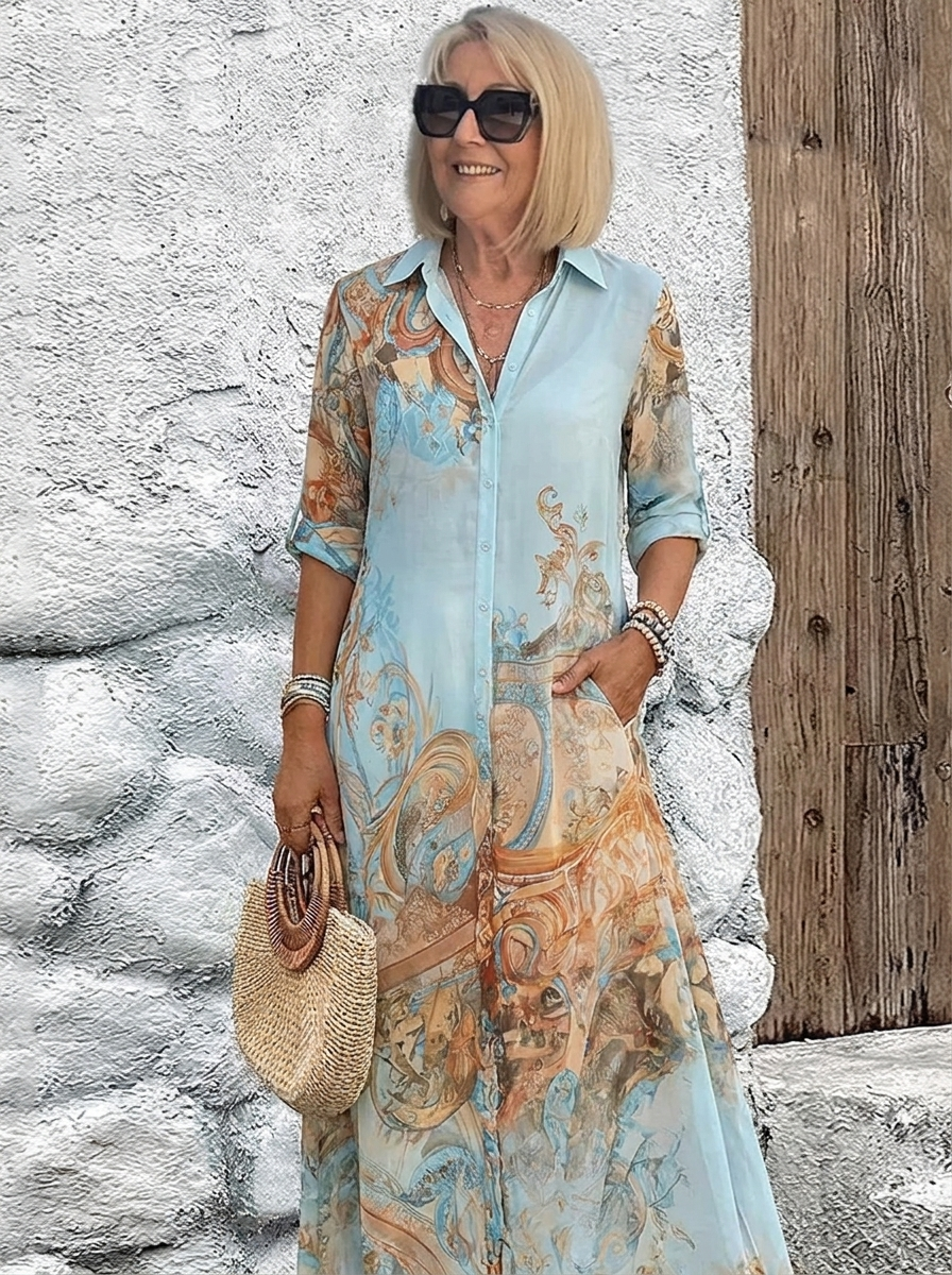 Woman in sunglasses wears the light blue button-front maxi dress with orange baroque print and rolled sleeves, holding a woven straw handbag. 1