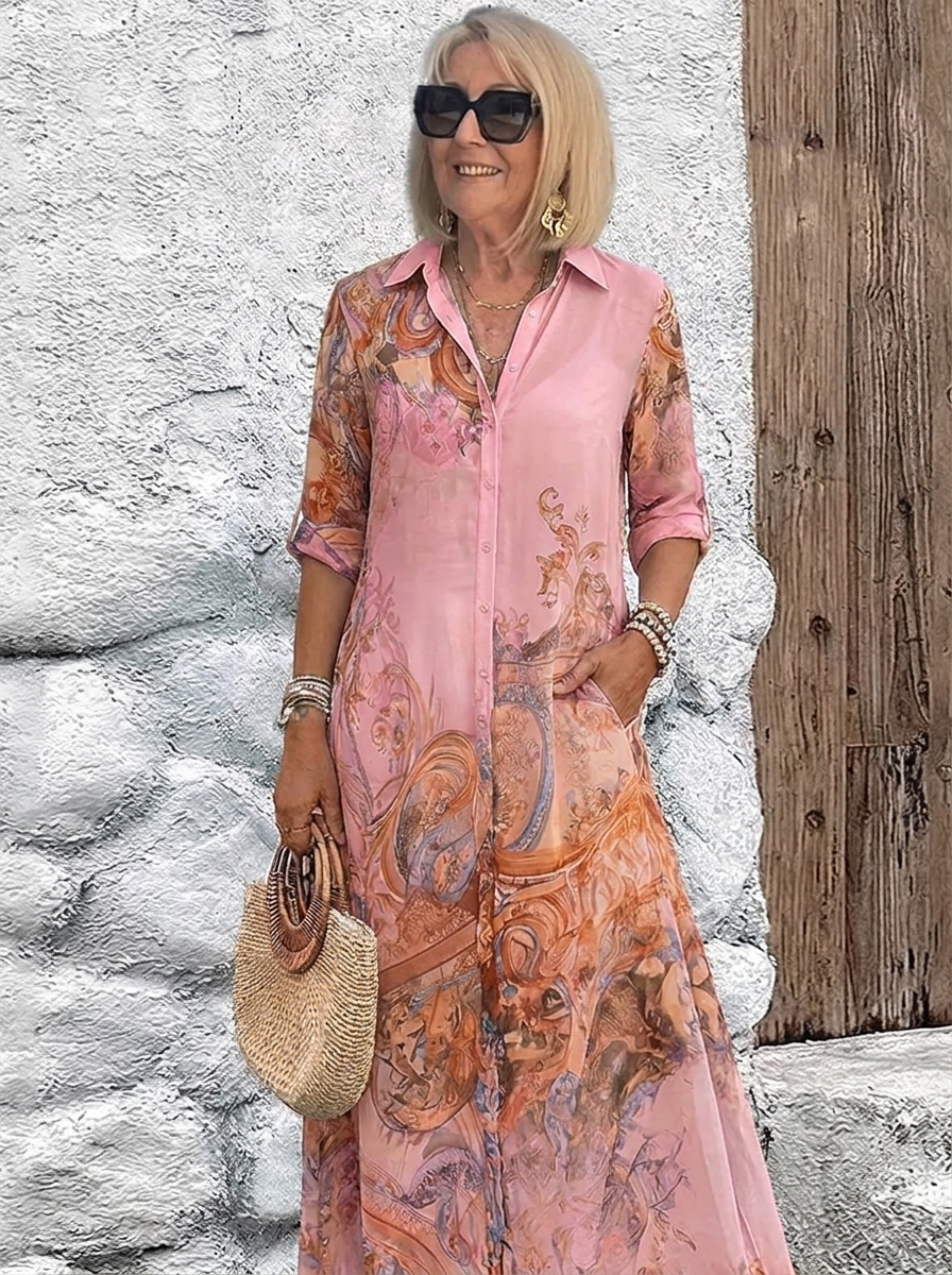 Model wears the soft pink version of the collared maxi dress with orange baroque print, one hand in a pocket, holding a straw bag. 4