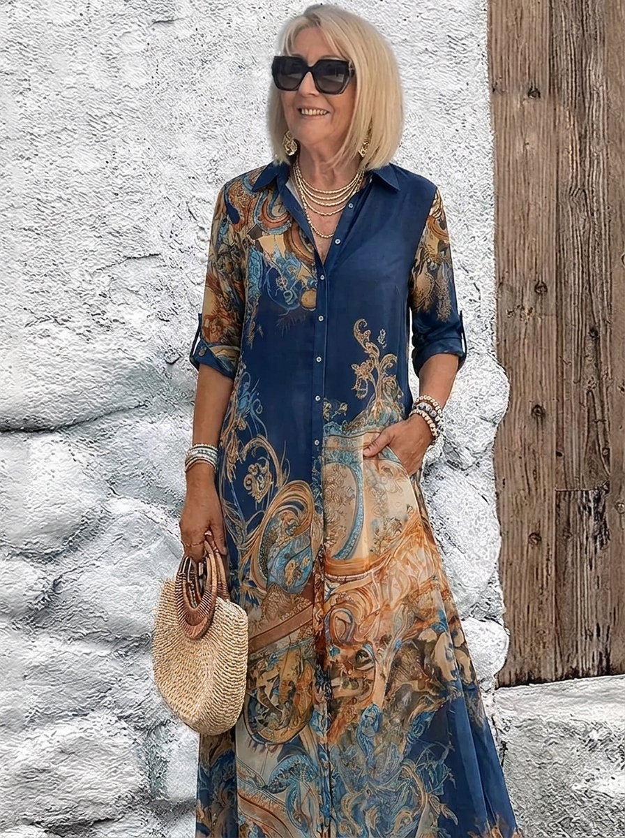 Same button-front maxi dress in deep navy blue with gold and orange baroque print, worn with layered necklaces and a round straw bag. 2