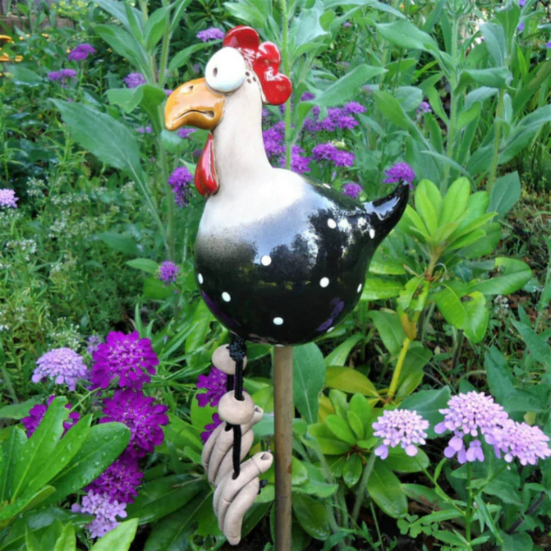 Black ceramic chicken with white polka dots and a white neck on a garden stake, dangling clay feet, among purple flowers. 2