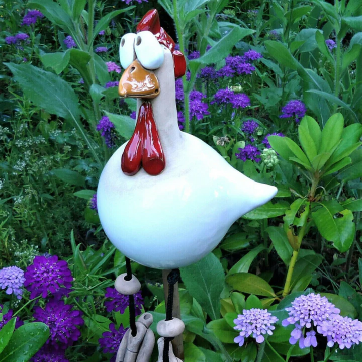 Glossy white ceramic chicken with bulging eyes and a red wattle on a garden stake, surrounded by purple blossoms. 3