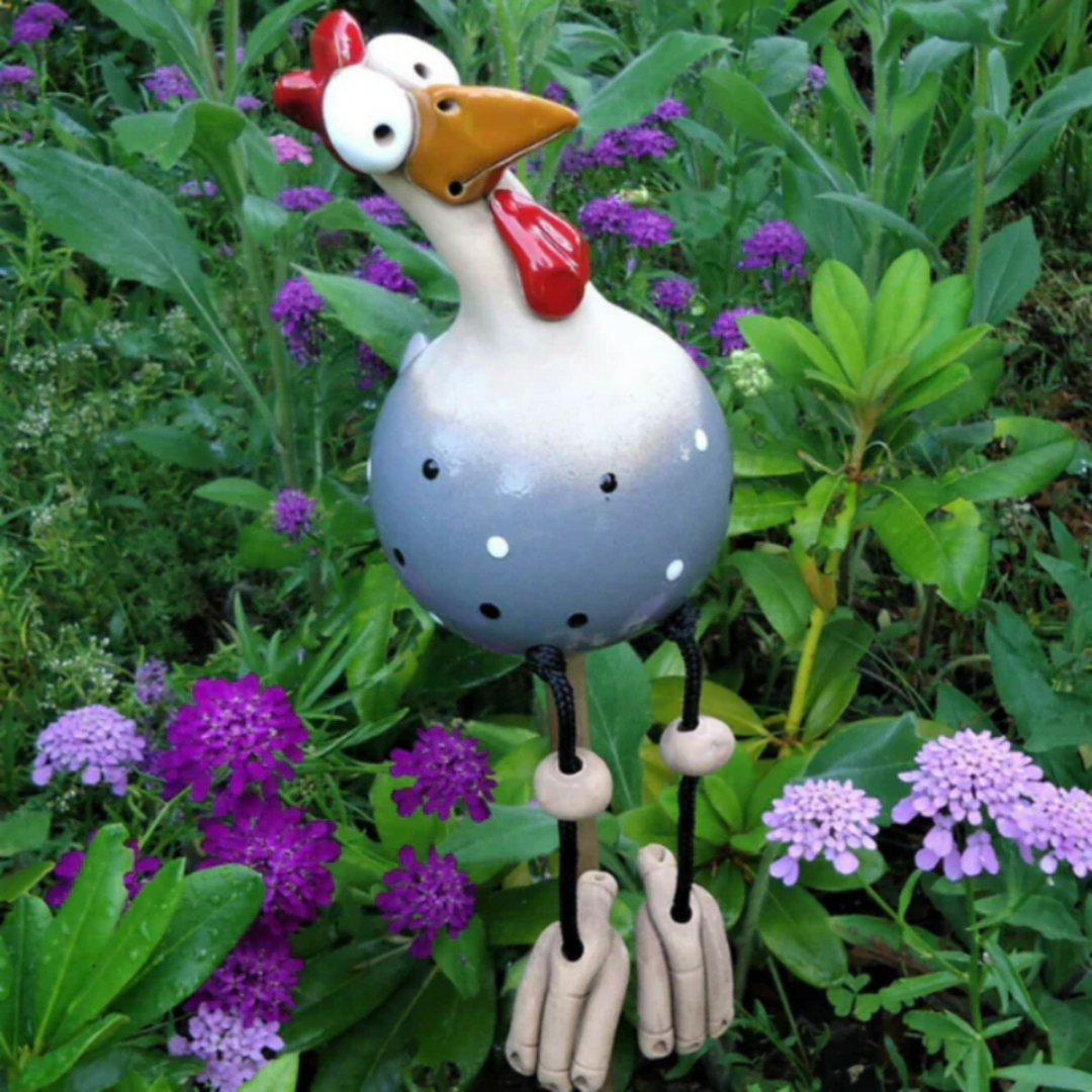 Gray polka-dot ceramic chicken with a long neck and a red wattle, black cord legs with clay beads and feet, among flowers. 5