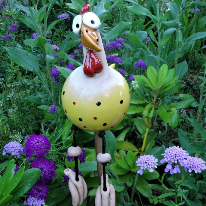 Yellow ceramic chicken with black and red dots and a red wattle on a garden stake, cord legs hanging, beside purple flowers. 6