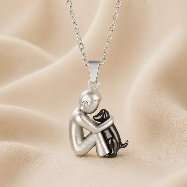 Side-view pendant of a matte silver-tone figure hugging a glossy black dog, hanging on a chain over cream fabric. 7