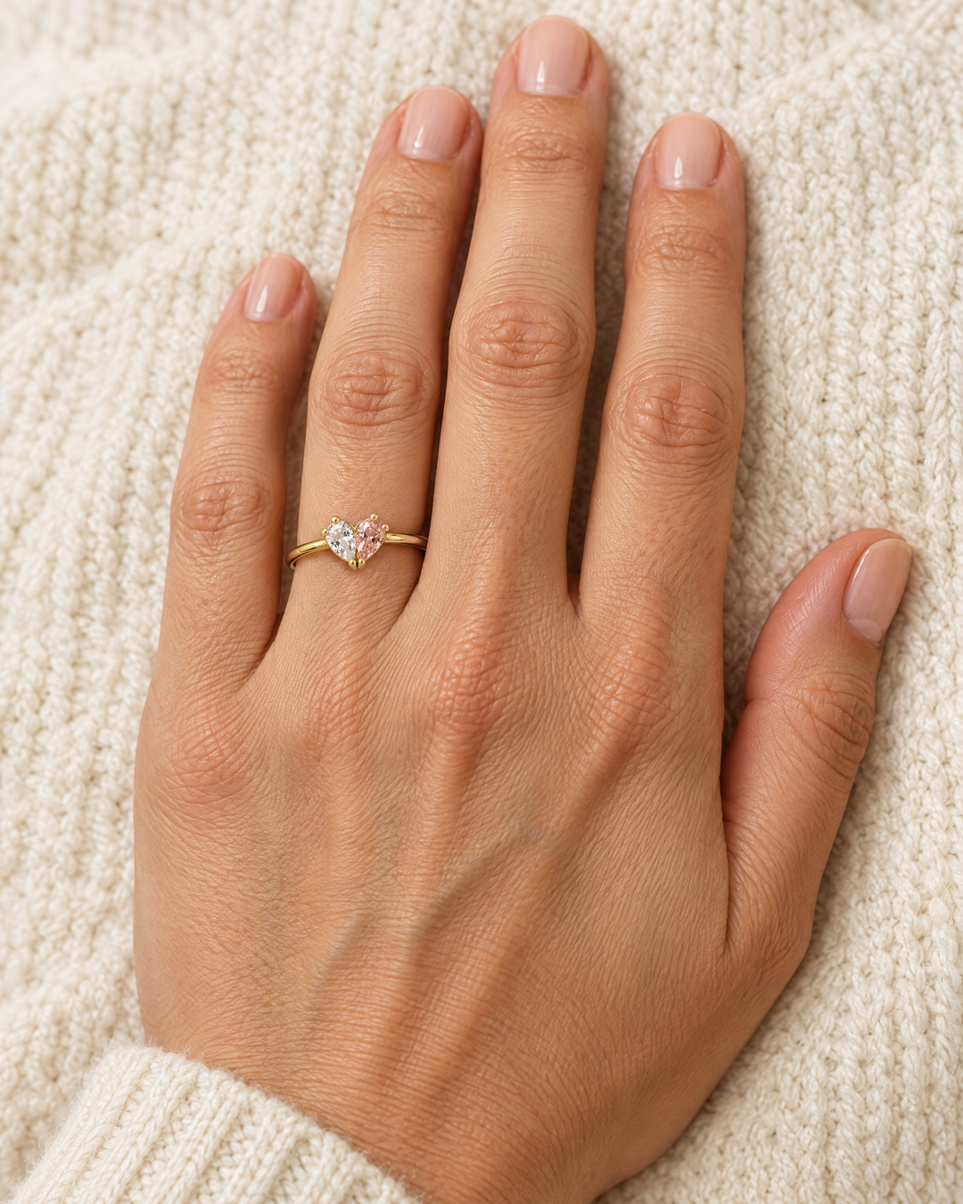 detail of the gold-toned heart ring worn on a hand with a clear and a pink teardrop stone.