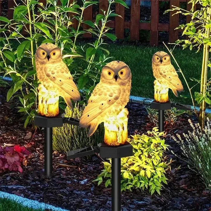 Three owl lights with golden-brown plumage glowing among green shrubs beside a wooden fence.