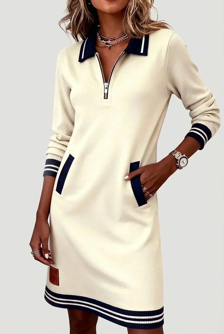 Model wearing the cream version of the dress, with navy striped collar, ribbed pockets and cuffs, one hand in a pocket. 2