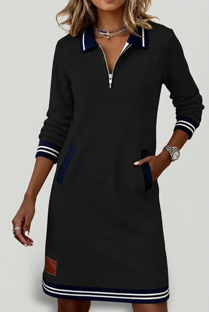 Black long-sleeve dress on a model with hand in pocket, showing navy striped collar, cuffs, hem and brown patch. 11