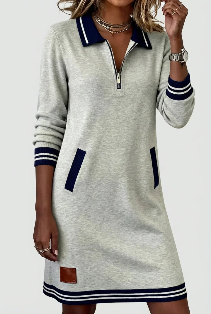 Model in heather gray long-sleeve zip dress with navy striped collar, cuffs and hem, one hand raised to her necklace. 1
