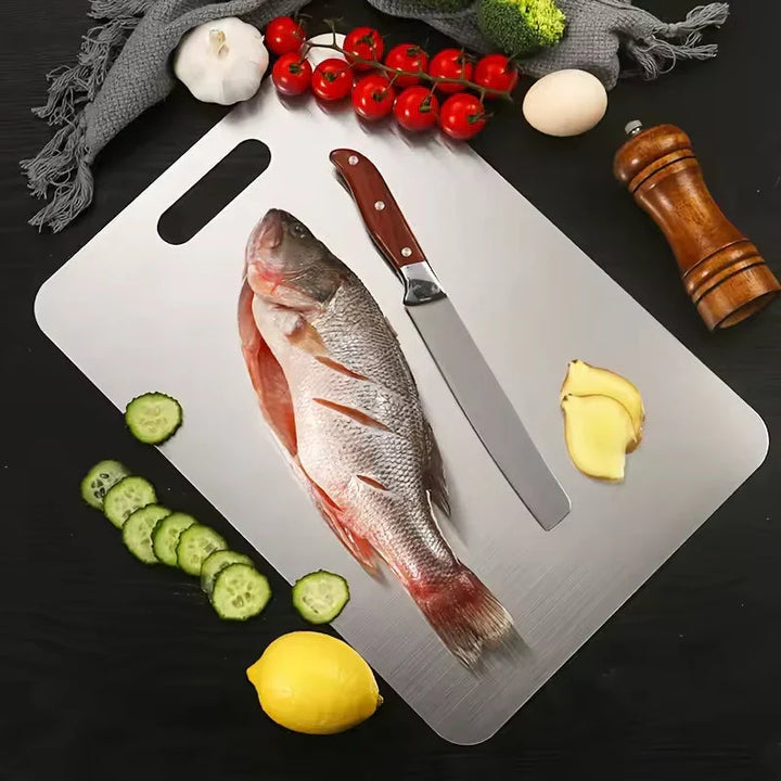 Overhead view of a metal cutting board holding a whole fish and a knife, surrounded by cucumber slices, lemon, ginger, tomatoes and garlic.